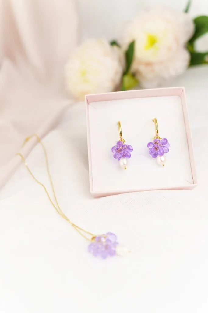 Kuvassa vaaleanpunainen korurasia jonka sisällä liilat helmikorvakorut kultaisilla korvakorurenkailla, taustalla vaaleanpunainen kangas ja kukkia