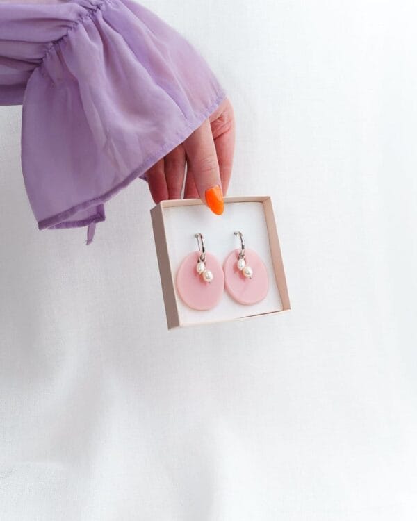 Kädessä korurasia jossa vaaleanpunaiset soikean muotoiset korvakorut helmillä ja hopeisilla korvakorurenkailla