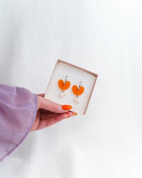 Kädessä korurasia jossa oranssit sydämen muotoiset korvakorut helmillä ja hopeisilla korvakorurenkailla