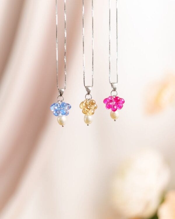 Kolme kaulakorua erivärisillä helmillä ja hopeisilla ketjuilla roikkuu telineessä, taustalla kukkia