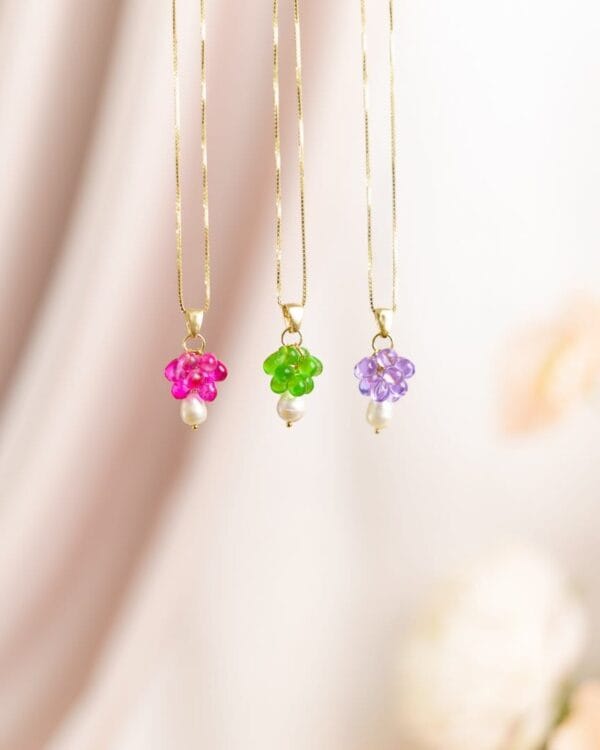Kolme kaulakorua erivärisillä helmillä ja kultaisilla ketjuilla roikkuu telineessä, taustalla kukkia