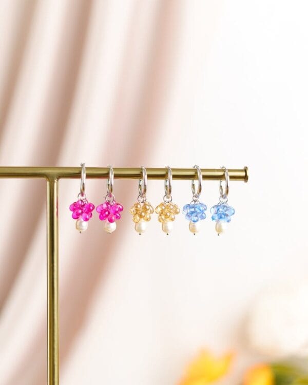 Kolmet korvakorut erivärisillä helmillä ja hopeisilla korvakorurenkailla roikkuu telineessä, taustalla kukkia