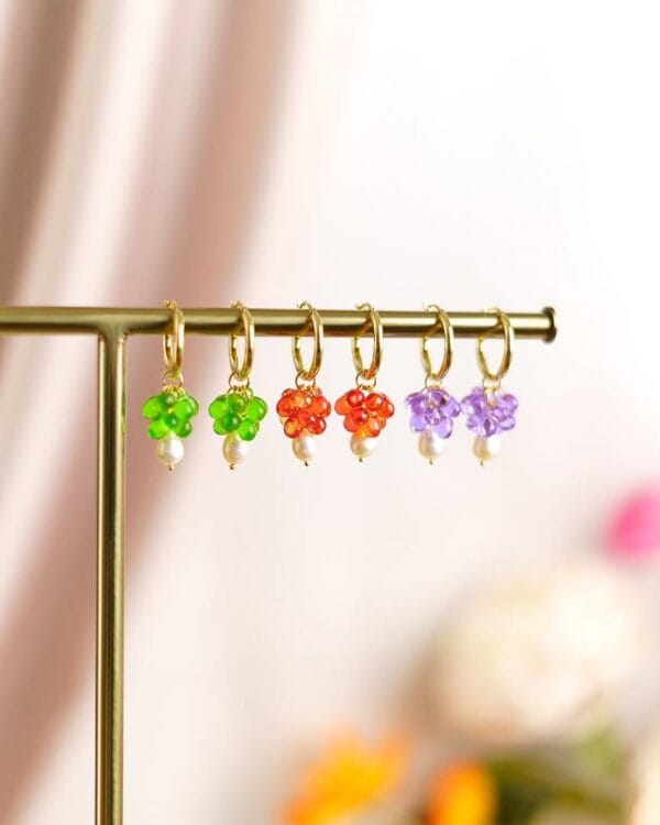 Kolmet korvakorut erivärisillä helmillä ja kultaisilla korvakorurenkailla roikkuu telineessä, taustalla kukkia