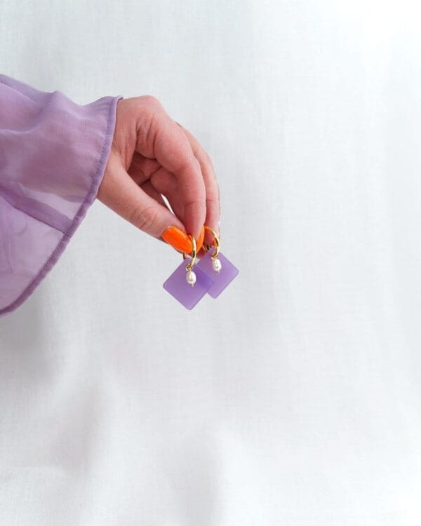 Kädessä neliön muotoiset ja liilan väriset korvakorut joissa helmi ja kultaiset korvakorurenkaat