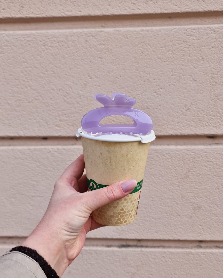 Kädessä take away -kahvikuppi jonka päällä vaalean liila hiusklipsi.