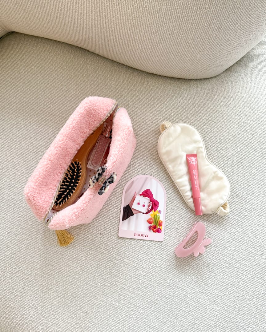 Kuvassa vaalealla sohvalla vaaleanpunainen meikkilaukku jossa kosmetiikkatarvikkeita sekä kaksi hiusklipsiä.